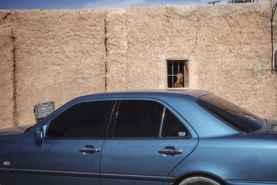 A hand gestures from a window behind a blue Mercedes-Benz parked against an adobe wall. The vibrant, metallic colors of the imported European cars often provide a sharp contrast to the monochrome ochre tones of the desert architecture and landscape.